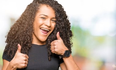 Genç güzel kadın kıvırcık saçlı başarı işareti el ile pozitif jest yapıyor başparmak gülümseyen ve mutlu. Neşeli bir ifadeyle, kazanan jest kameraya bakıyor.