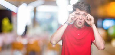 İzole arka planda kırmızı tişört giyen genç yakışıklı adam parmaklar, Uykulu ve sabah yorgunluk için yorgun gözleri açmaya çalışıyor