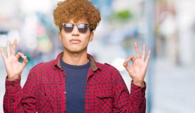 Kapalı giyen güneş gözlüğü rahatla afro saçlı yakışıklı delikanlı ve gülümseyen gözlerle parmak ile meditasyon jest yapıyor. Yoga kavramı.