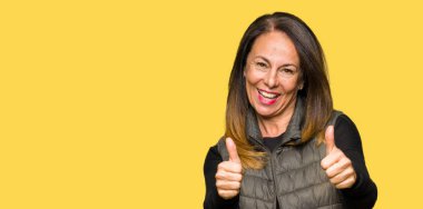 Kış onaylama yelek giyen güzel orta yaş kadın el ile pozitif jest yapıyor gülümseyen ve başarı için mutlu Yaşasın. Kamera, kazanan jest arıyorsunuz.