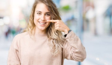 Güzel sarışın genç kadın giyen sweatershirt izole üzerinden yüz ve burun, neşeli gülümseyerek el parmak ile işaret arka plan