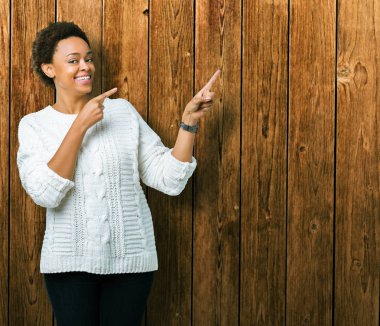 Gülümseyen ve iki elleri ve parmakları ile tarafına işaret kamera bakarak izole arka plan üzerinde kazak giyen güzel genç Afro-Amerikan kadın.