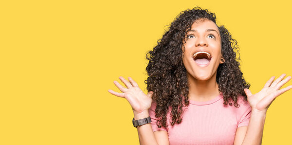 Young beautiful woman with curly hair wearing pink t-shirt celebrating crazy and amazed for success with arms raised and open eyes screaming excited. Winner concept