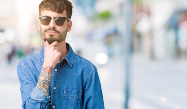 Genç yakışıklı adam çene düşünme hakkında soru, dalgın ifade üzerine el ile izole arka plan üzerinde güneş gözlüğü takıyor. Düşünceli yüzüyle gülümsüyor. Şüphe kavramı.