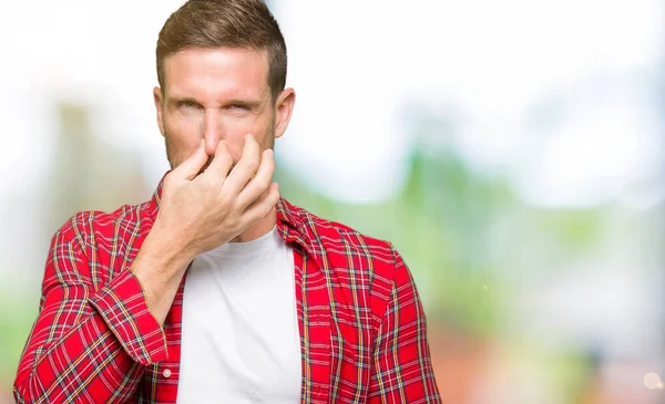 Handsome man wearing casual shirt smelling something stinky and disgusting, intolerable smell, holding breath with fingers on nose. Bad smells concept.