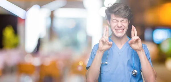 若い医師は 希望と目を閉じて指を横切って笑顔で孤立した背景の上に医療の制服を着て 幸運と迷信的概念 — ストック写真