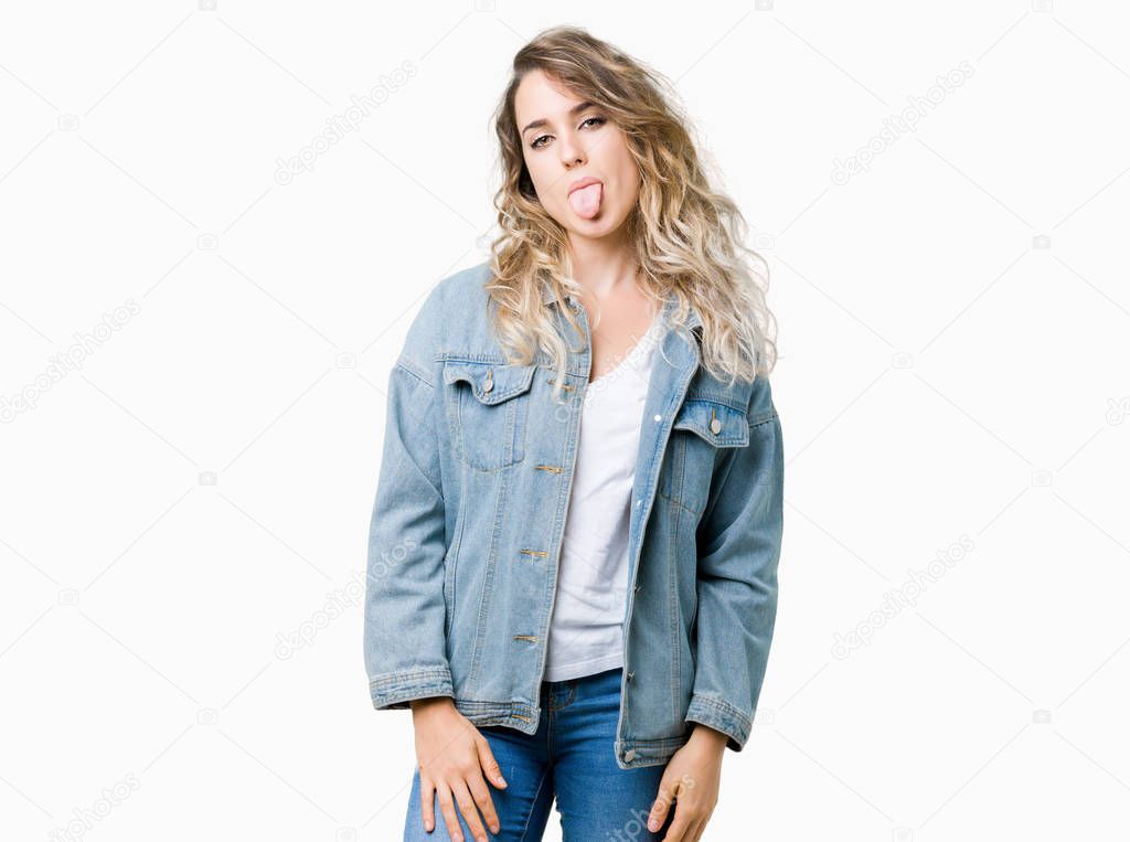 Beautiful young blonde woman wearing denim jacket over isolated background sticking tongue out happy with funny expression. Emotion concept.