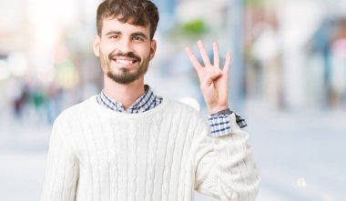 Gösterilen arka plan üzerinde kış kazak giyen genç yakışıklı adam izole ve parmakları ile işaret kendinden emin ve mutlu gülümseyerek süre dört numara.