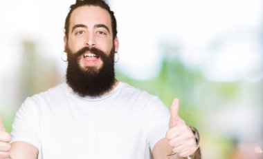 Uzun saç ve sakal el ile olumlu hareketi yaparken rahat beyaz t-shirt başarı işareti giyen genç hipster adamla başparmak gülümseyen ve mutlu. Neşeli bir ifadeyle, kazanan jest kameraya bakıyor.