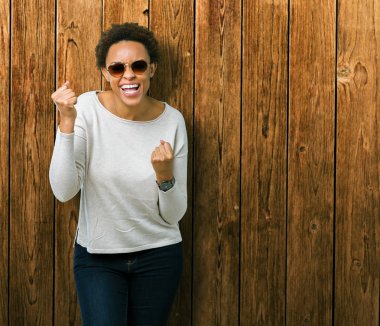 Güzel genç Afro-Amerikan kadın üzerinde güneş gözlüğü takıyor arka plan çok mutlu ve heyecanlı kaldırdı, silah ile kazanan jest yapıyor gülümseyen ve başarı için çığlık izole. Kutlama kavramı.