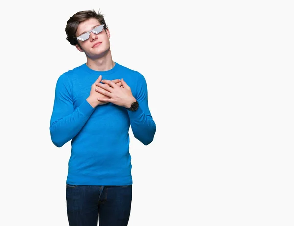 Young man wearing funny thug life glasses over isolated background smiling with hands on chest with closed eyes and grateful gesture on face. Health concept.