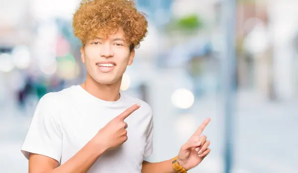 カジュアルな白いTシャツを着てアフロヘアの若いハンサムな男は微笑み 両手と指を横に向けてカメラを見ている — ストック写真