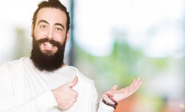 Mutlu ve neşeli gülümseyen genç hipster adamla uzun saç ve sakal sportif kazak gösteren palm el giyen ve, baş parmakları Tamam jest yapıyor