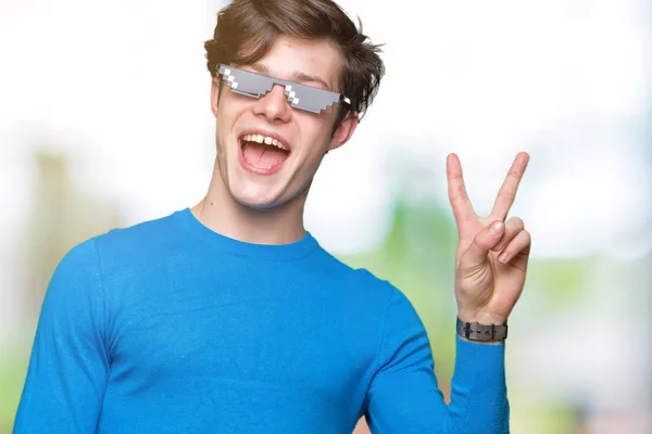 Young man wearing funny thug life glasses over isolated background smiling with happy face winking at the camera doing victory sign. Number two.