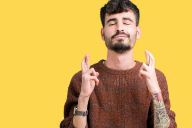 Geçiş parmakları umut ve gözleri kapalı ile gülümseyen izole arka plan üzerinde kış kazak giyen genç yakışıklı adam. Şans ve batıl kavramı.