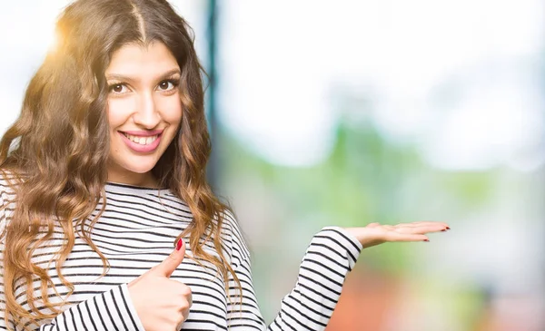 Çizgili kazak giyen genç güzel kadın palmiye el gösteren ve başparmak ile ok jest yapıyor, mutlu ve neşeli gülümseyen