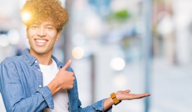 Afro saçlı kot giyen genç yakışıklı adam ceket gösteren palm el ve yukarıya, mutlu ve neşeli gülümseyen baş parmakları Tamam jest yapıyor