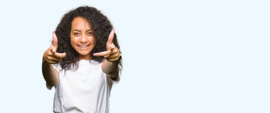 Kıvırcık saçlı olumlu ve neşeli gülümseyen casual beyaz t-shirt işaretleme ve parmaklar, kamerayla giyen genç güzel kız