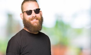 Genç hipster adam yüzünde mutlu ve güzel bir gülümseme ile güneş gözlüğü takıyor. Şanslı kişi.