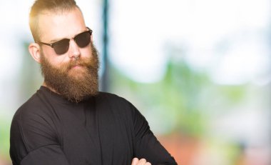 Çapraz kolları olan yüzündeki güneş gözlüğü şüpheci ve sinir, onaylamayan ifade giyen genç hipster adam. Negatif kişi.