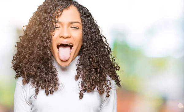 Young beautiful woman with curly hair wearing turtleneck sweater sticking tongue out happy with funny expression. Emotion concept.