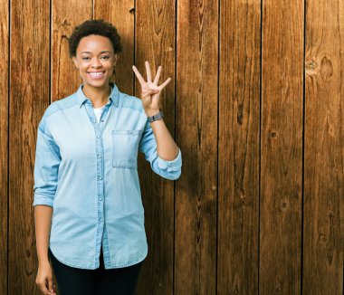 İzole arka plan üzerinde genç güzel Afrikalı Amerikalı kadın gösteren ve parmak sayısı dört ile işaret kadar güven ve mutlu gülümseyen.