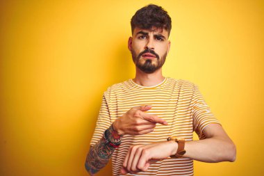 Dövme ile genç adam izole sarı arka plan üzerinde ayakta çizgili t-shirt dikiliyor acele seyretmek zaman, sabırsızlık, üzgün ve son gecikme için kızgın işaret