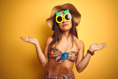 Kadın tatile bikini ve ananas güneş gözlüğü üzerinde izole sarı arka plan clueless ve silah ve eller ile karıştı ifade kaldırdı. Şüphe kavramı.