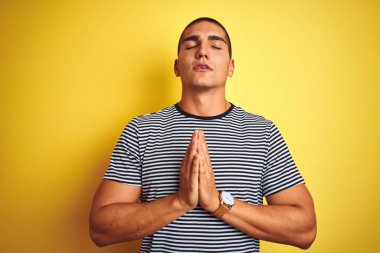 Genç yakışıklı adam sarı izole arka plan üzerinde çizgili t-shirt giyen yalvarıyor ve çok duygusal ve endişeli yüzünde umut ifadesi ile birlikte elleri ile dua. Af dilemek istiyorum. Din kavramı.