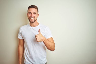 Hareketi el ile mutlu yaşasın yapıyor izole arka plan üzerinde rahat beyaz t-shirt giyen genç yakışıklı adam. Başarı gösteren kameraya bakarak ifade onaylama.