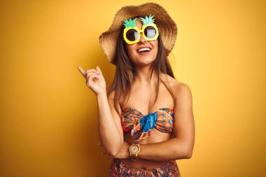 Yüz üzerinde büyük bir gülümseme ile izole sarı arka plan üzerinde bikini ve ananas güneş gözlüğü giyen tatil kadın, kamera bakarak yan el ve parmak ile işaret.