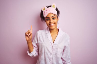 Genç Afrikalı Amerikalı kadın pijama ve Maske giyen izole pembe arka plan gösteren ve parmak bir numara ile işaret kadar emin ve mutlu gülümseyen.