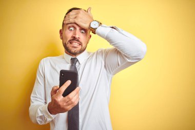 Sarı izole arka plan üzerinde Smartphone kullanarak genç yakışıklı iş adamı baş el ile vurguladı, utanç ve sürpriz yüz ile şok, öfkeli ve sinirli. Korku ve hata için üzgün.