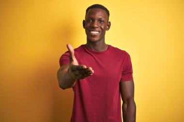 Genç Afrikalı Amerikan adam izole sarı arka plan üzerinde ayakta kırmızı t-shirt giymiş dostu tebrik ve karşılama olarak el sıkışması sunuyor. Başarılı iş.
