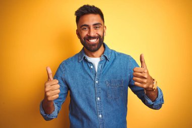 Genç Hint adam giyen denim gömlek ayakta izole sarı arka plan başarı işareti ile olumlu jest yapıyor el, thumbs up gülümseyen ve mutlu. Neşeli ifade ve kazanan jest.