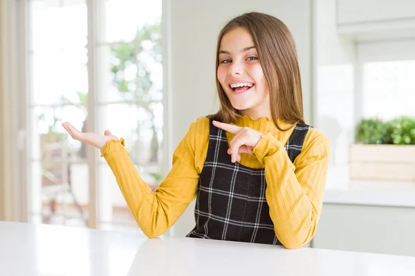 年轻美丽的金发女孩穿着休闲黄色毛衣在家里惊讶和微笑的相机 同时呈现与手和手指指点 — 图库照片