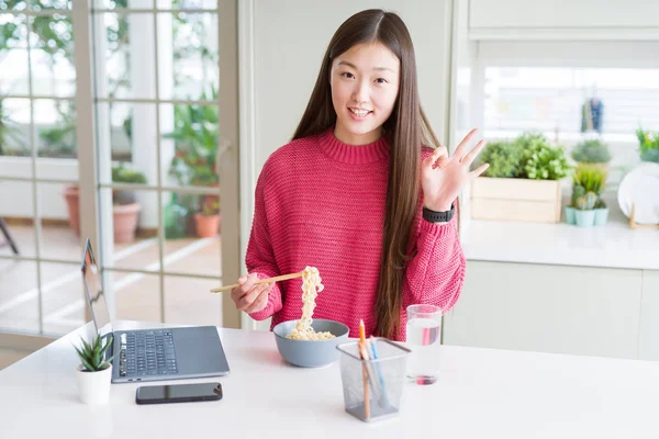 Güzel Asyalı kadın dizüstü bilgisayar kullanarak çalışan ve parmaklarıyla ok işareti yapıyor Asya noodle yeme, mükemmel sembolü