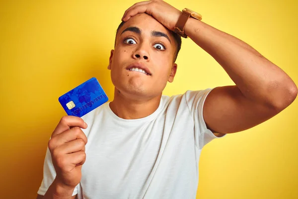 年轻的巴西客户男子拿着信用卡站在孤立的黄色背景强调与手在头上 震惊与羞愧和惊讶的脸 愤怒和沮丧 因错误而感到恐惧和不安 — 图库照片