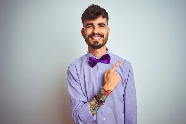 Young man with tattoo wearing purple shirt and bow tie over isolated white background cheerful with a smile of face pointing with hand and finger up to the side with happy and natural expression on face