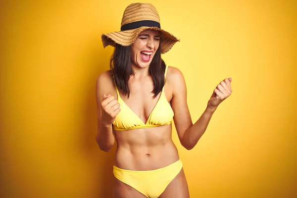 Beautiful woman wearing yellow bikini and summer hat over isolated yellow background very happy and excited doing winner gesture with arms raised, smiling and screaming for success. Celebration concept.