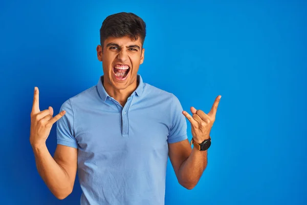 カジュアルなポロを着た若いインド人男性が孤立した青い背景の上に立ち 手を挙げてロックシンボルをする狂気の表情で叫ぶ 音楽スターだヘビーコンセプト — ストック写真
