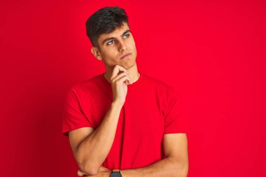 İzole kırmızı arka plan üzerinde duran tişört giyen genç hintli adam çenesi üzerinde soru, dalgın ifade yi düşünüyor. Düşünceli bir yüzle gülümsüyor. Şüphe kavramı.