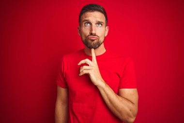 Kırmızı izole arka planda rahat t-shirt giyen genç yakışıklı adam çene üzerinde parmak ile şüphe hakkında konsantre düşünme ve merak bakıyor