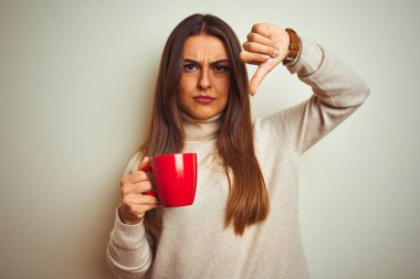Genç güzel kadın kızgın yüzü ile izole beyaz arka plan üzerinde duran kahve kırmızı fincan içme, negatif işareti başparmak aşağı ile sevmediğim gösteren, ret kavramı