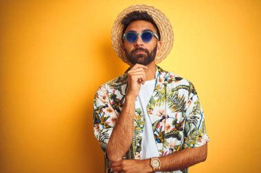 Tatil Hint adam üzerinde el ile izole sarı arka plan üzerinde çiçek gömlek şapka güneş gözlüğü giyiyor soru hakkında düşünme, dalgın ifadesi. Düşünceli bir yüz ile gülümsüyor. Şüphe kavramı.