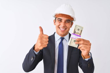 Genç yakışıklı mimar adam izole beyaz arka plan üzerinde büyük bir gülümseme ok işareti yapıyor mutlu dolar tutan kask giyen, parmakları ile başparmak, mükemmel işareti