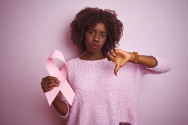 Kızgın yüz ile izole pembe arka plan üzerinde ayakta kanser şerit duran genç Afrikalı Afro kadın, aşağı başparmak ile nefret gösteren negatif işareti, ret kavramı