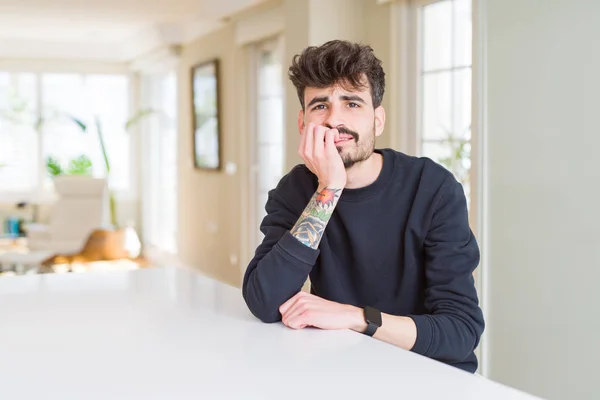 stock image Young man wearing casual sweatshirt sitting on white table looking stressed and nervous with hands on mouth biting nails. Anxiety problem.