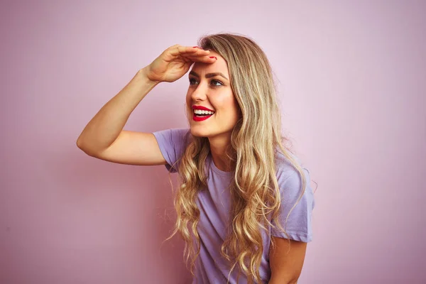 Young beautiful woman wearing purple t-shirt standing over pink isolated background very happy and smiling looking far away with hand over head. Searching concept.