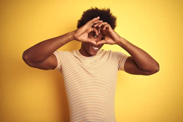 American man with afro hair wearing striped t-shirt standing over isolated yellow background Doing heart shape with hand and fingers smiling looking through sign